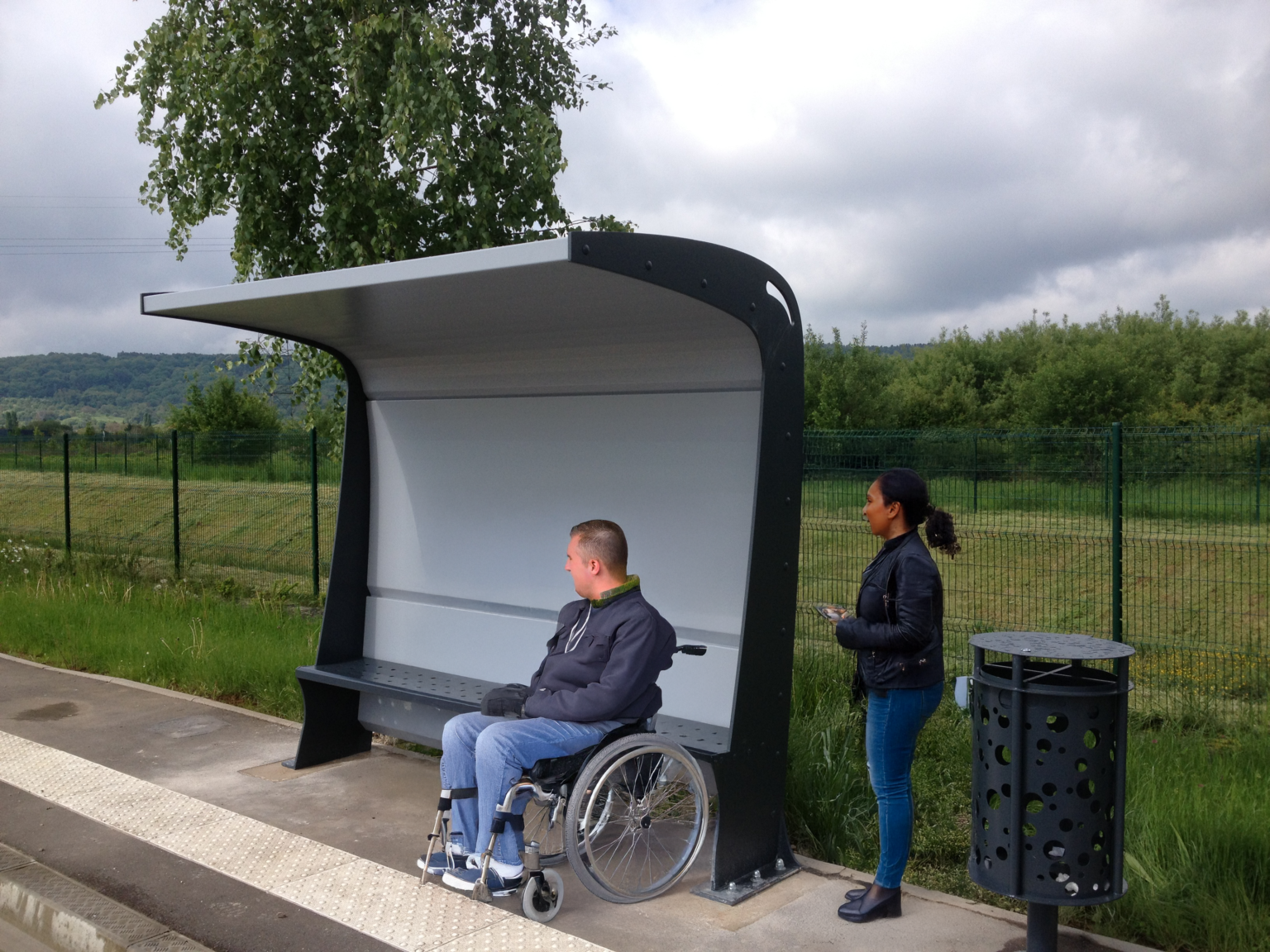Un homme en fauteuil roulant et une femme debout attendent à un abribus moderne et accessible, conçu en acier. L’abribus offre une protection contre les intempéries avec son toit incurvé et un banc intégré. L’environnement est verdoyant, avec un fond de collines et un ciel nuageux. Une poubelle au design assorti est placée à proximité, soulignant une approche soignée du mobilier urbain inclusif